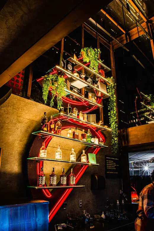 Bar do Amargot: estante iluminada com garrafas e plantas, em pé-direito duplo