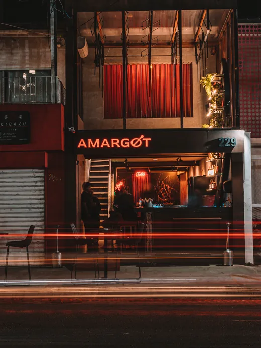 Fachada do bar Amargot à noite: letreiro de neon vermelho, número 229 e mesas na calçada, com a rua iluminada