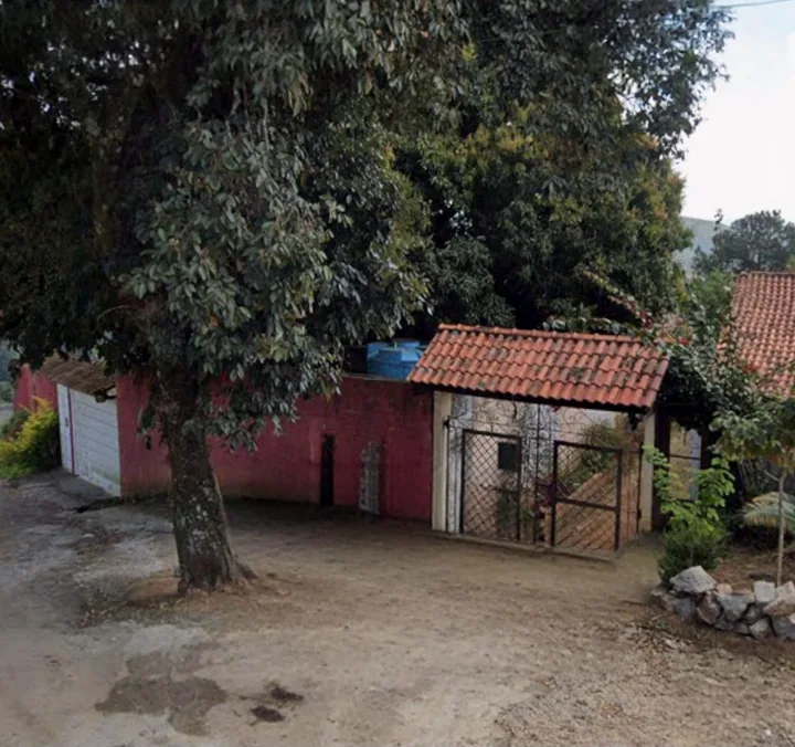 Portão de entrada do local: muro vermelho e telhadinho de telhas ao fim de uma rua de terra em subida, com árvore e montanha ao fundo